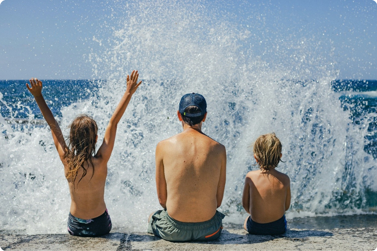 Das Bild zeigt drei Kinder, die mit dem Rücken zur Kamera auf einer niedrigen Steinmauer oder Uferkante sitzen. Sie blicken auf das Meer.